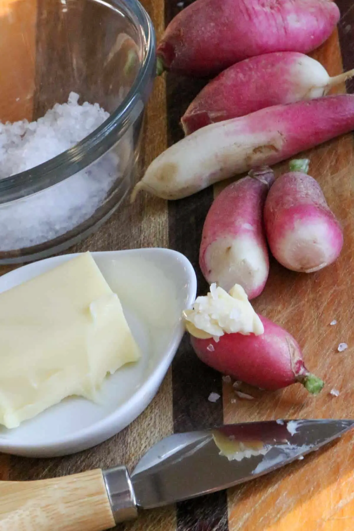 Several French breakfast radishes on a wooden cutting board with a glass bowl with sea salt in the background, a white dish with butter on the left side, and a cheese knife with wooden handle in the foreground.