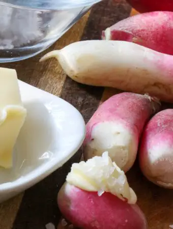 French breakfast radishes on a wooden board one radish has butter and salt on i there is a white dish with butter to the left of the radishes and a glass dish with coarse salt at the top left.