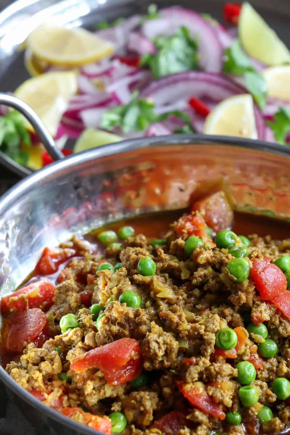 Lamb mince, peas, onion, and diced tomatoes in a rich sauce in a silver balti bowl, with a salad in the background consisting of sliced red onion, wedges of lemons and limes, sliced chilies and cilantro on a silver plate.