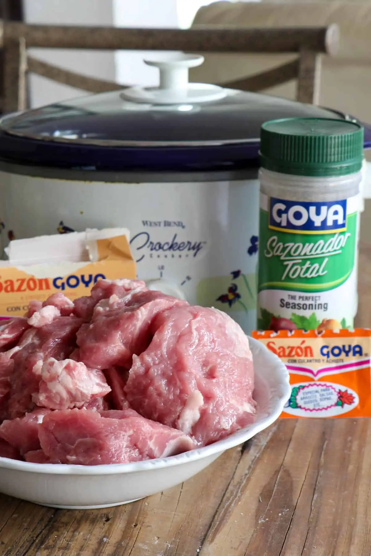 A crockpot, a white bowl filled with uncooked pork meat cuts, a box of Sazon Goya and packet of Sazon Goya Seasoning, and bottle of Goya Sazonador Total seasoning.