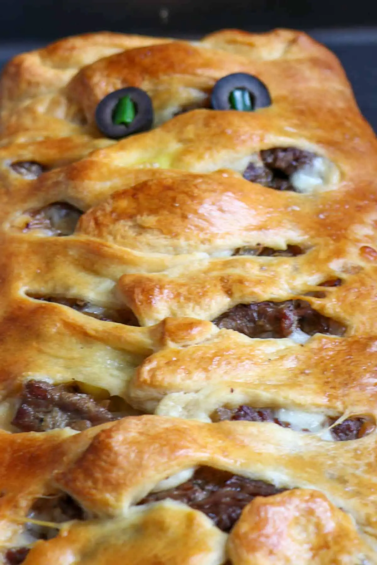 Sliced rib eye steak wrapped with cooked crescent roll dough that has been cut into 1 inch intervals and placed on top of the sliced meat to resemble mummy wrappings and eyes made of black olives with green onions as pupils.