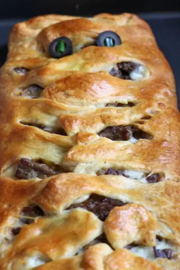 Sliced rib eye steak wrapped with cooked crescent roll dough that has been cut into 1 inch intervals and placed on top of the sliced meat to resemble mummy wrappings and eyes made of black olives with green onions as pupils.