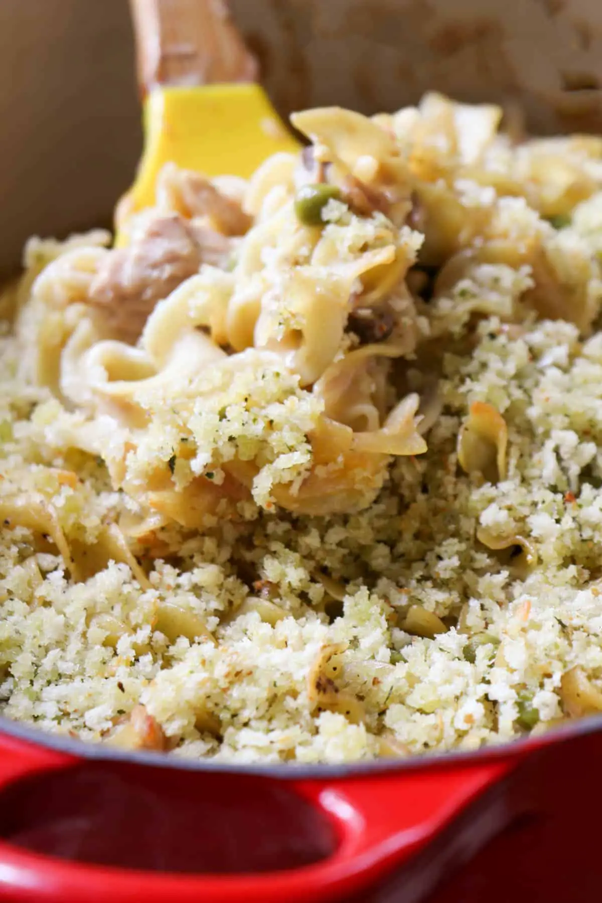 Noodles cooked with tuna and peas in a creamy sauce with a panko topping and a yellow spoon cradling some of the tuna noodle casserole which is in a red handled Dutch oven.