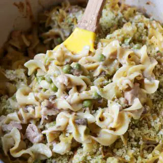 Noodles cooked with tuna and peas in a creamy sauce with a panko topping and a yellow spoon cradling some of the tuna noodle casserole.