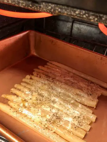 White asparagus topped with melting cheese, butter and seasonings in a roasting tray placed under a broiler.