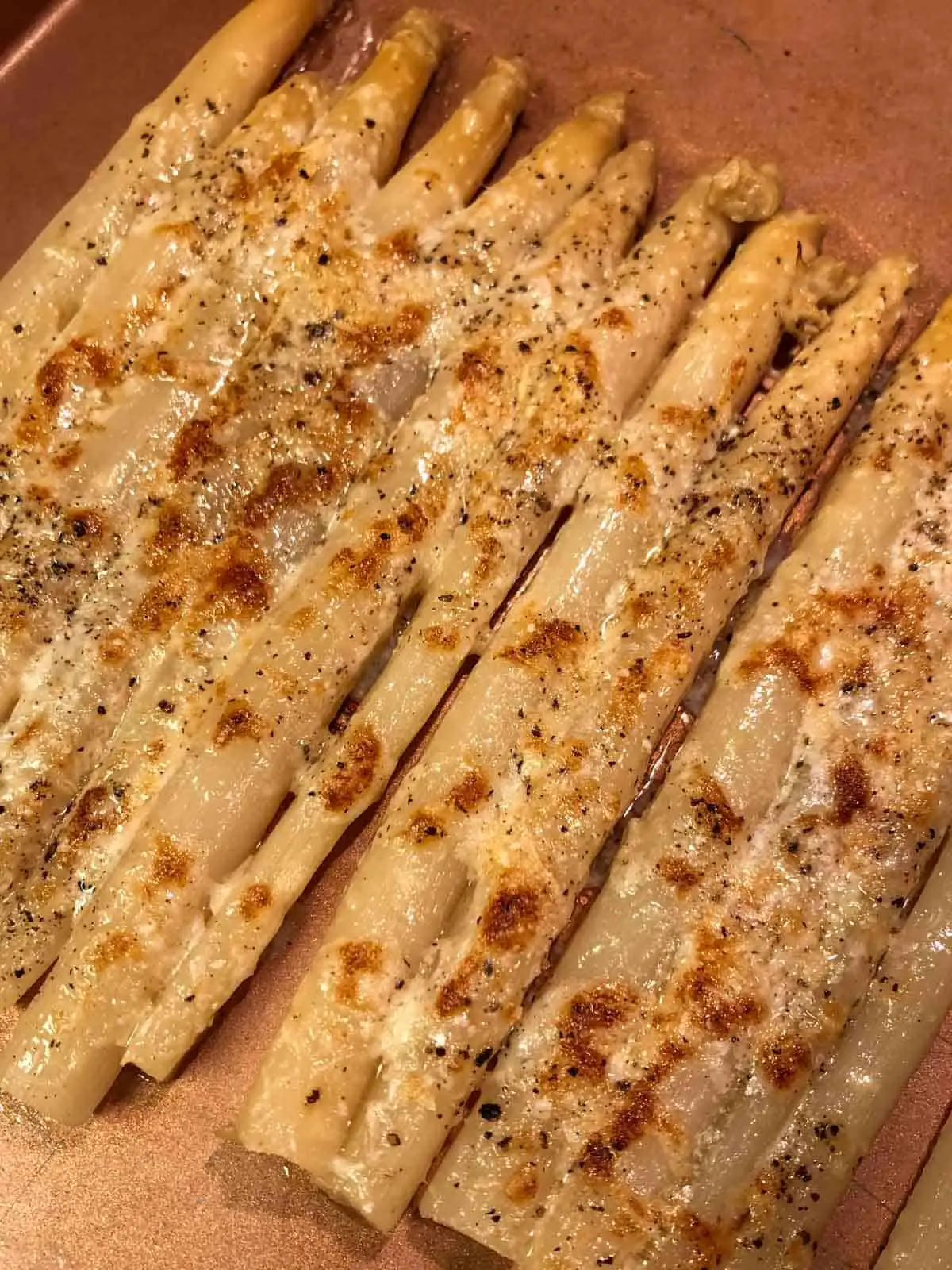 White asparagus in a roasting tray which was broiled and has browned melted cheese on top.
