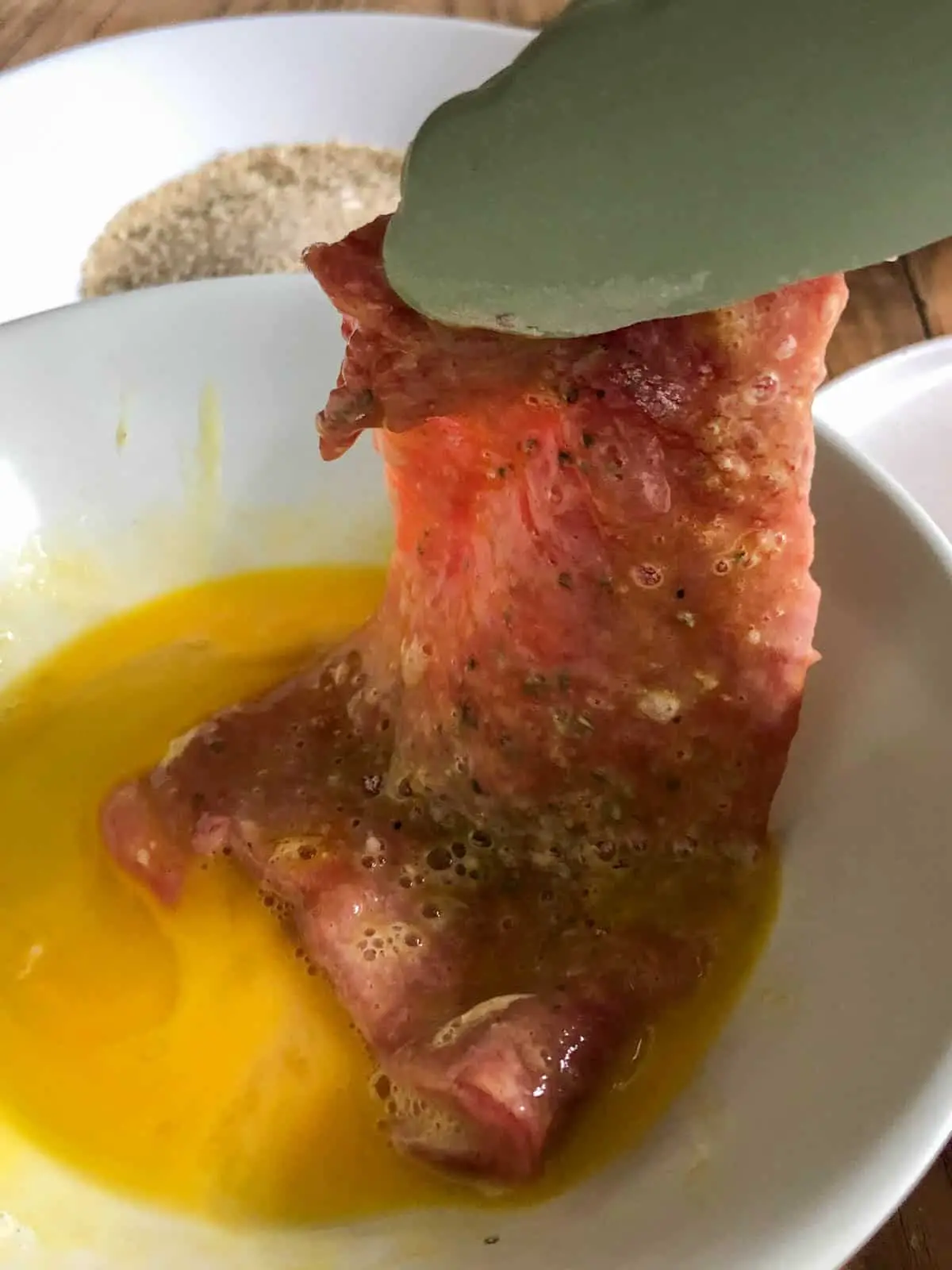 Veal cutlet that has been dipped in egg in a white bowl with a pair of blue tongs holding up the veal cutlet and a white bowl filled with bread crumbs in the background.