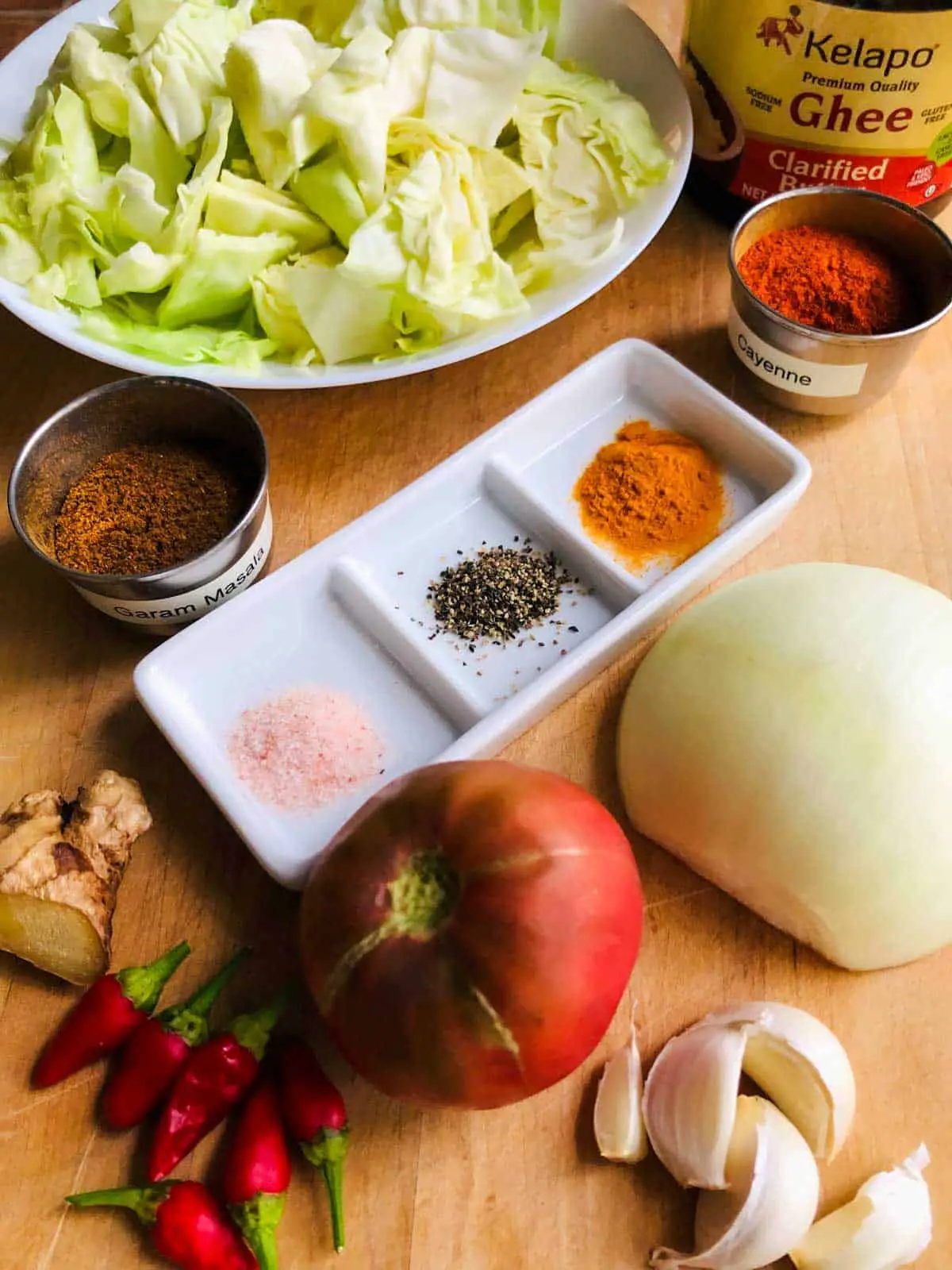 Chopped cabbage in a white bowl, ghee, cayenne and garam masala in silver bowls. salt, pepper, and turmeric in a white dish, ginger, several red chilies, tomato, half and onion, and several garlic cloves on a wooden board.
