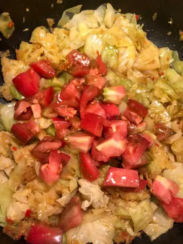 Cooked cabbage with seasonings and garlic, ginger, chilies, and onions and chopped tomato on top of the cabbage in a large skillet.