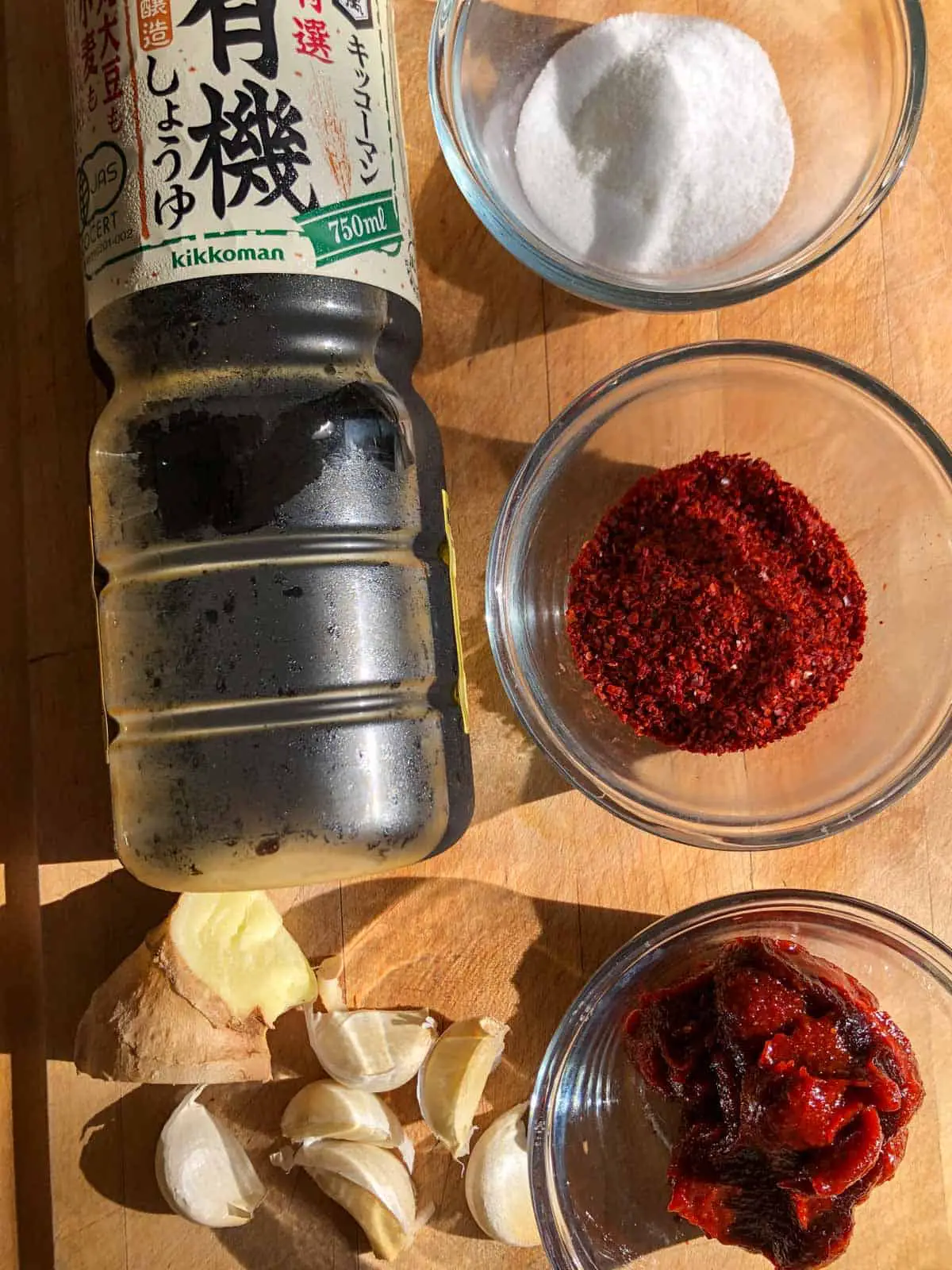 Bottle of soy sauce, glass dishes containing sugar, gochugaru, gochujang, piece of ginger, and some garlic cloves on a wooden board.