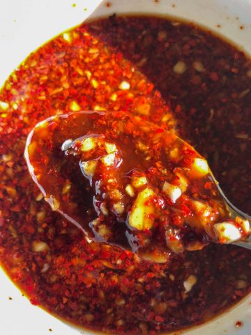 A spicy red sauce with garlic and chili flakes in a white bowl with a spoon containing some of the sauce in the foreground.