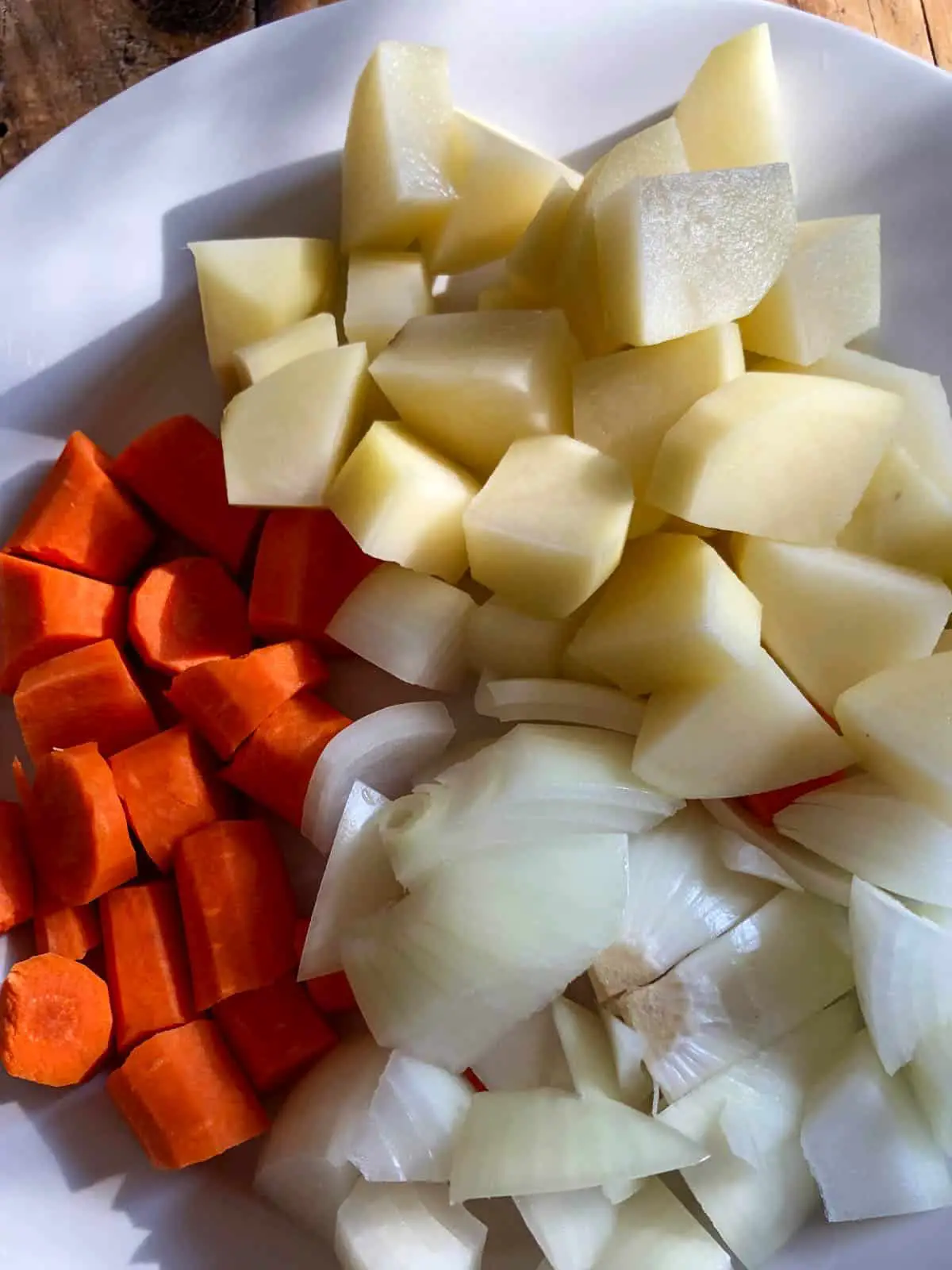 Cut up carrots, cut up potatoes, and cut up onions on a white plate.