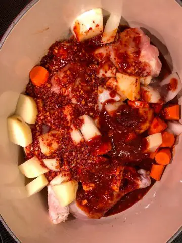 Chicken drumsticks, cut up carrots, onions, and potatoes in a red spicy sauce in a dutch oven.