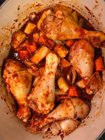 Chicken drumsticks, cut up carrots, onions, and potatoes in a red spicy sauce in a dutch oven.