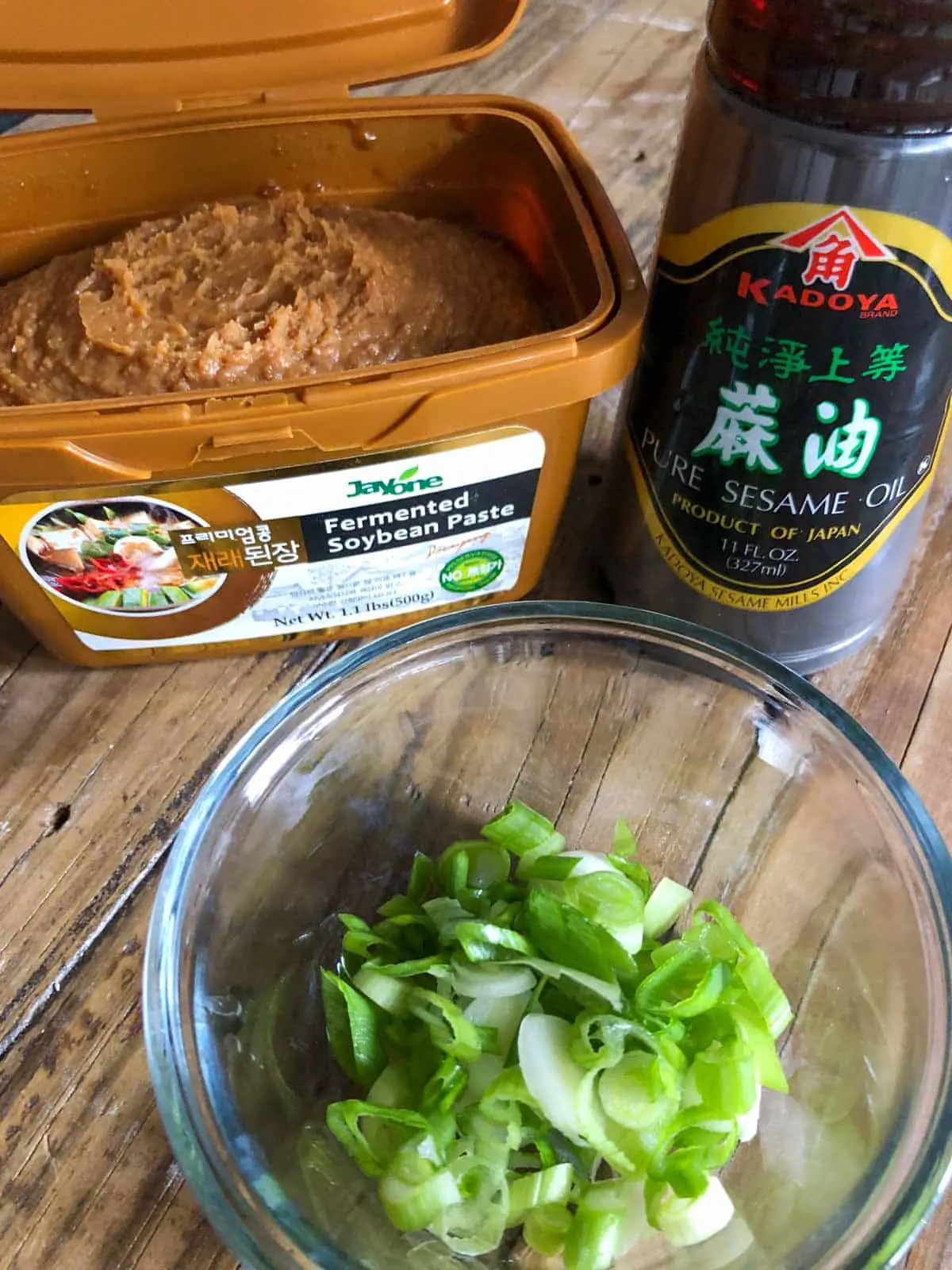 A container of fermented soybean paste, a bottle of sesame oil, and a glass bowl with sliced green onions.