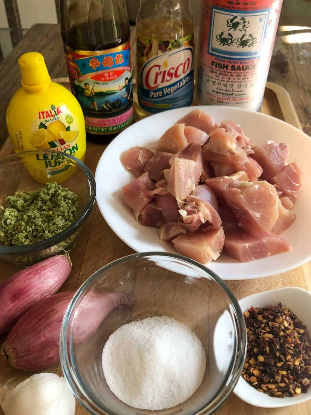 Pieces of chicken thighs cut up in a white dish, red chili flakes in a white dish, sugar in a glass bowl, garlic, 2 shallots, a glass dish with frozen lemongrass, a bottle of lemon juice, bottle of oyster sauce, vegetable oil, and a bottle of fish sauce.