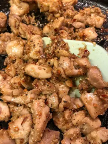 Pieces of chicken that have been cooked with lemongrass and other spices in a cast iron skillet with a blue silicone utensil.