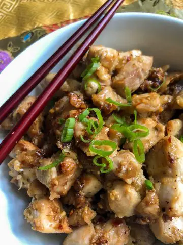 Pieces of chicken thigh that were cooked with lemongrass, garlic, chili flakes and shallots, garnished with green onion in a white bowl with chopsticks resting on the side.