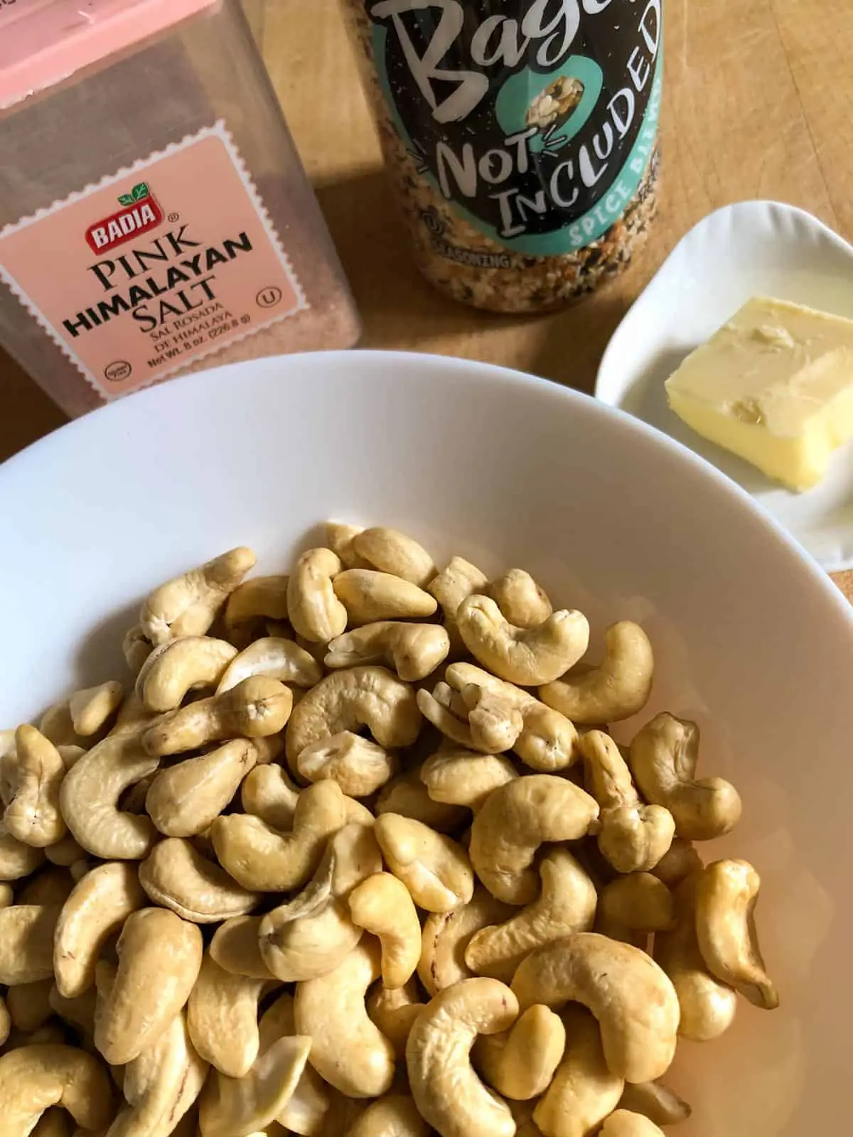 Cashews in a white bowl, container of Pink Himalayan Salt, bottle of Bagel not included seasoning, white dish with butter.