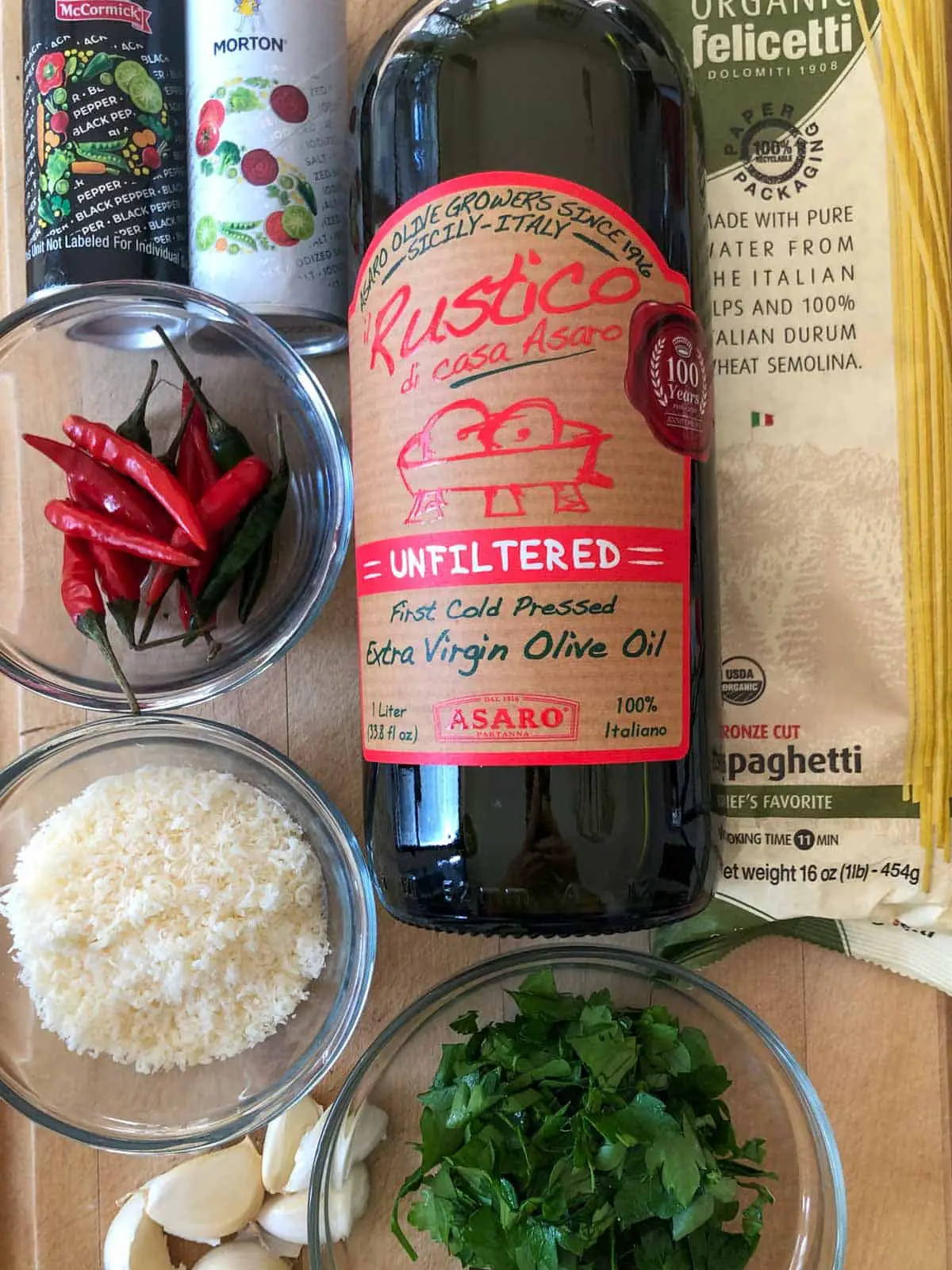 Salt and pepper shakers, a glass bowl with Thai chilies, a glass bowl with grated Parmesan cheese, garlic cloves, a glass bowl with chopped Italian parsley, a bottle of olive oil and a package of spaghetti.