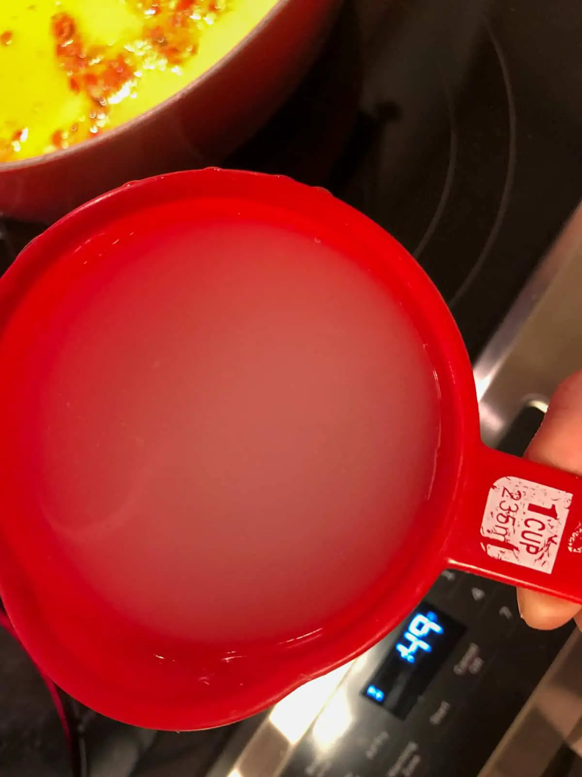 A red measuring cup containing starchy pasta water and a silver stove in the background.