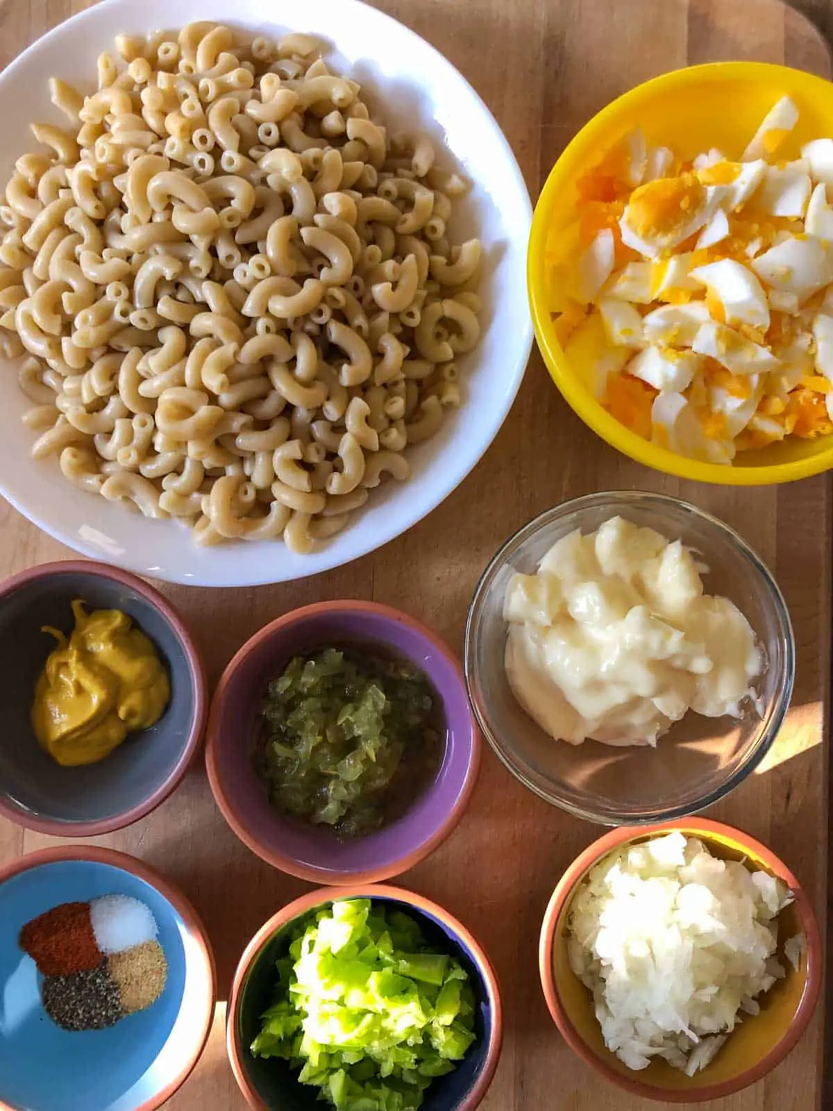 Cooked macaroni in a white dish, chopped hard boiled eggs in a yellow bowl, small bowls containing each of the following: mustard, sweet relish, mayonnaise, seasonings, chopped green bell pepper, and diced onion.