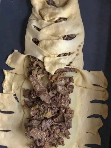 Sliced rib eye steak, diced onion, and cheese slices partially wrapped with crescent roll dough that has been cut into 1 inch intervals and placed on top of the sliced meat to resemble mummy wrappings.