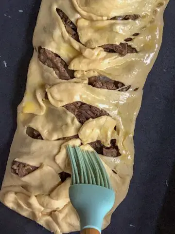 Sliced rib eye steak wrapped with crescent roll dough that has been cut into 1 inch intervals and placed on top of the sliced meat to resemble mummy wrappings with a blue baster basting egg wash on the dough.