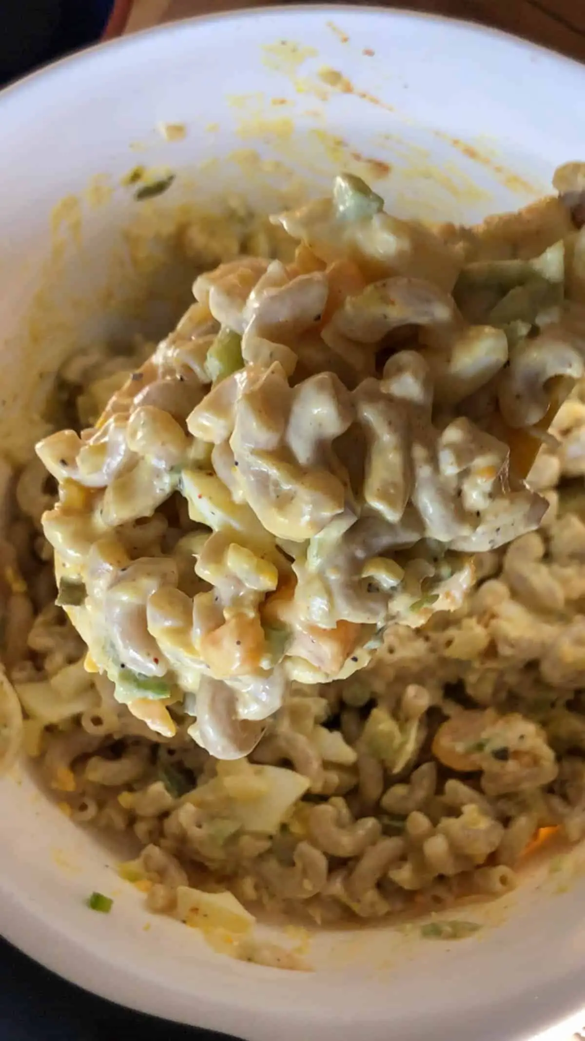 Macaroni, chopped hard boiled eggs, and chopped green peppers coated with mayonnaise and mustard in a white mixing bowl with a spoon containing this macaroni in the foreground.