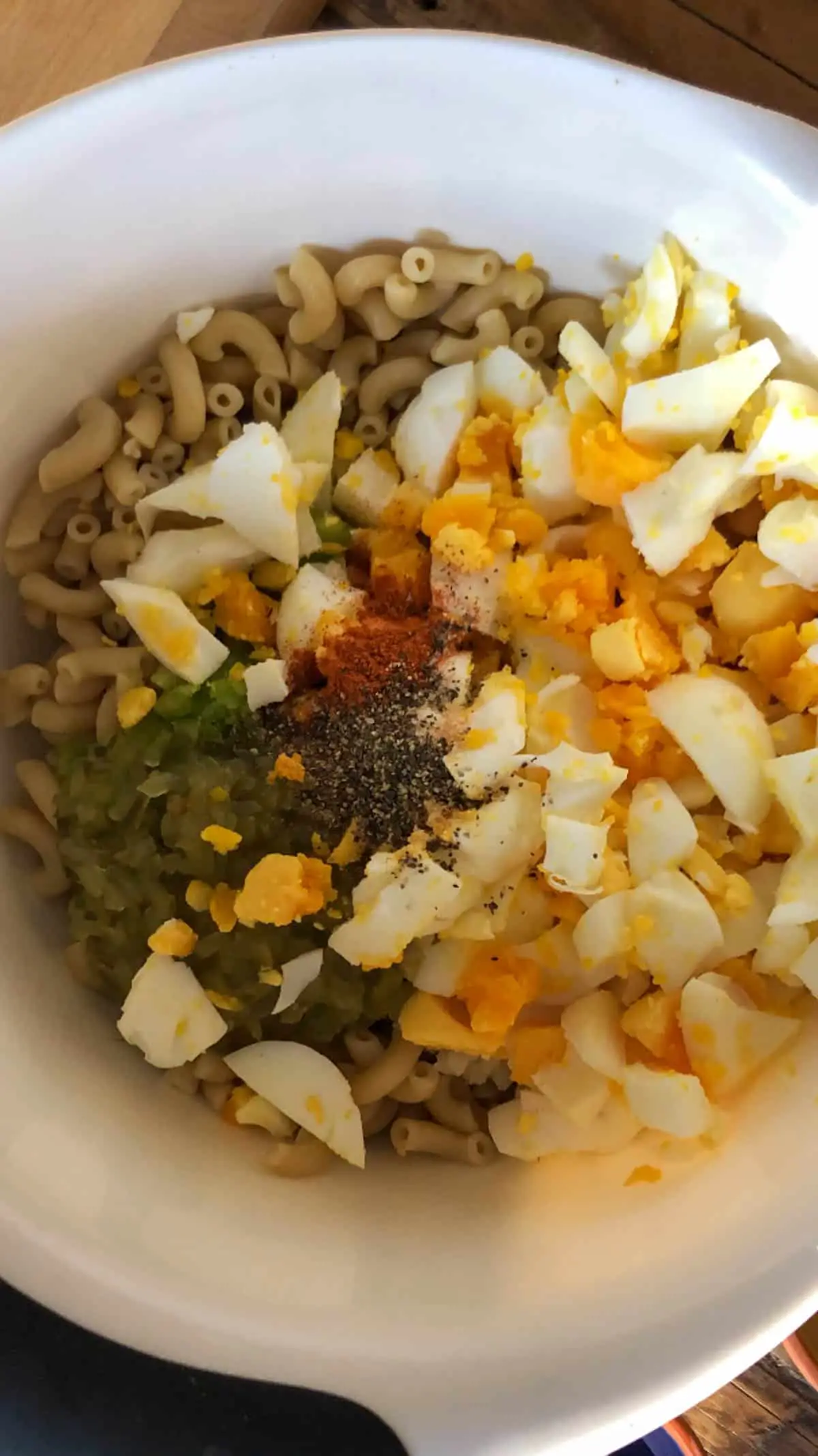 Cooked macaroni, chopped hard boiled eggs, seasonging, sweet relish and green bell pepper in a white mixing bowl.