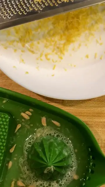 Lemon zest in a white bowl with a zester resting on top of the bowl and a green juicer with lemon seeds and lemon juice underneath the white bowl.