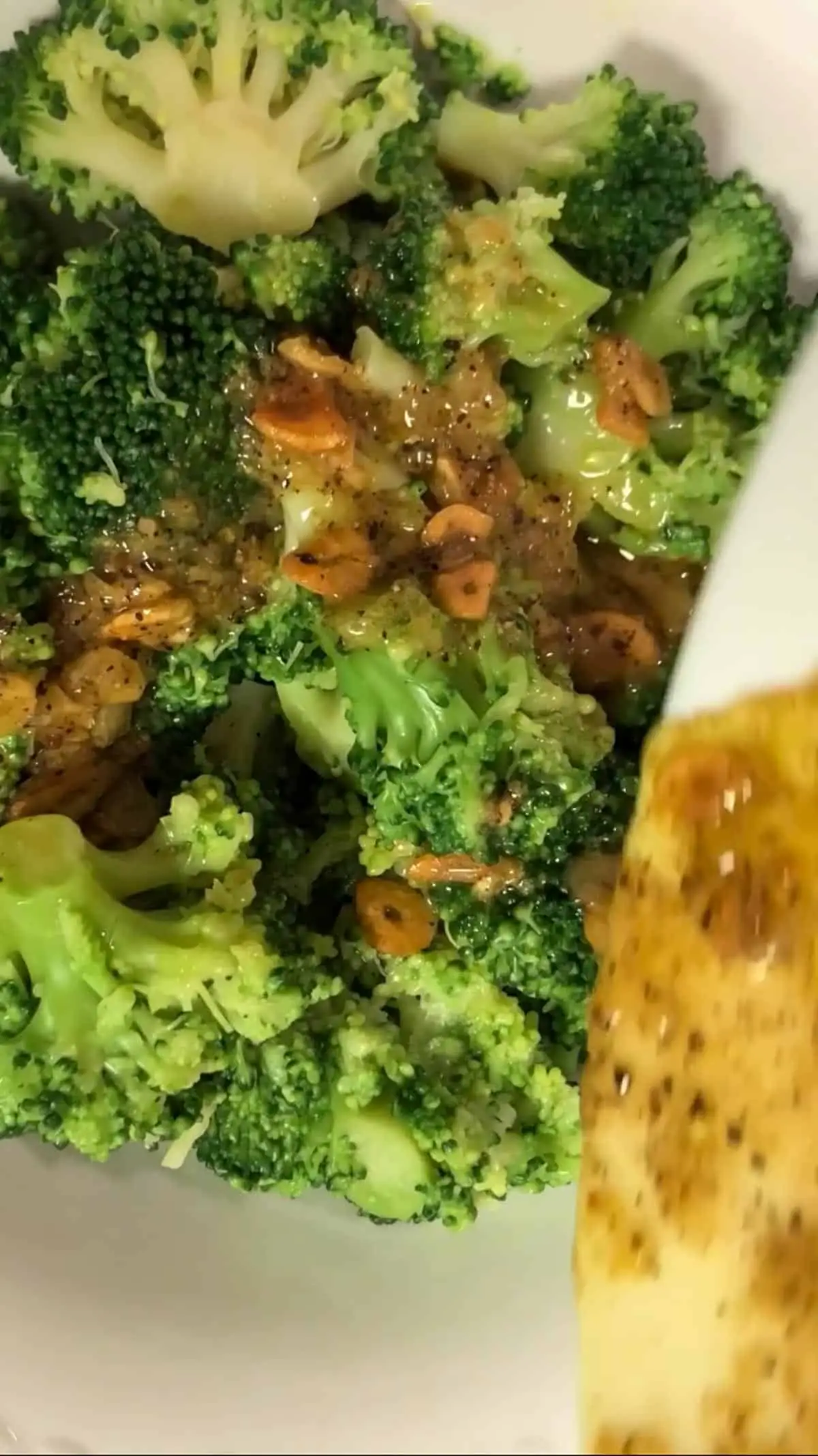 Broccoli florets in a white bowl with slivers of toasted garlic and a lemony sauce in a white bowl being drizzled on top of the garlic.