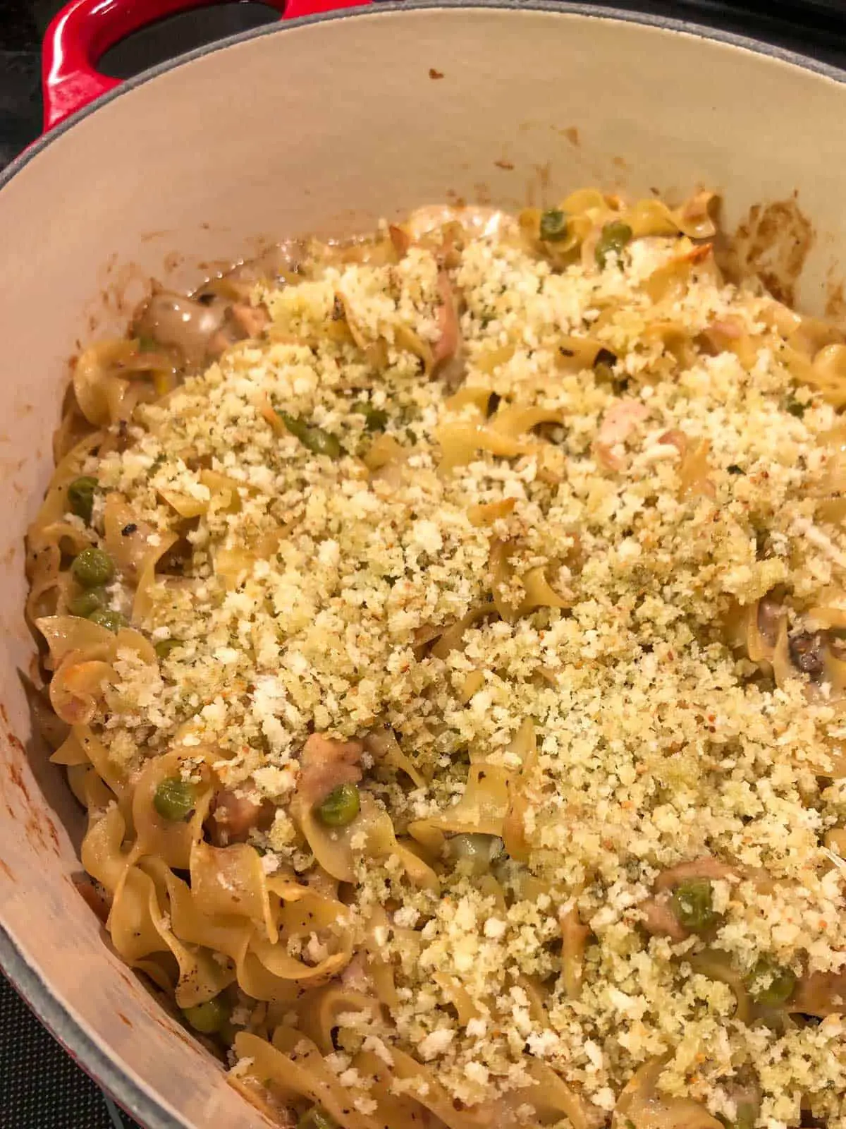 Noodles cooked with tuna and peas in a creamy sauce with a panko topping in a red handled Dutch oven.