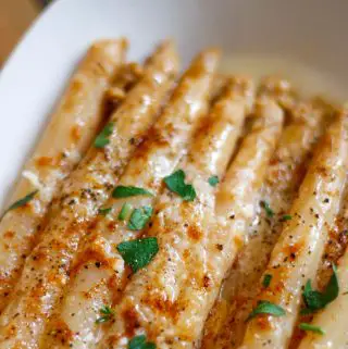 White asparagus in butter topped with browned melted cheese and garnished with Italian parsley in a white dish.