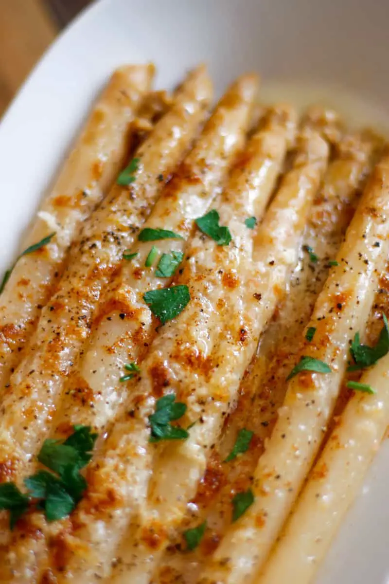 White asparagus in butter topped with browned melted cheese and garnished with Italian parsley in a white dish.