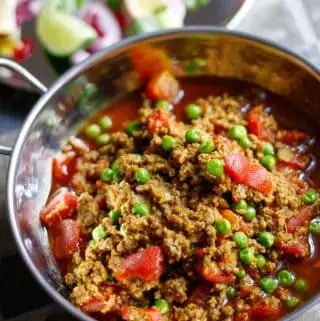 Lamb mince, peas, onion, and diced tomatoes in a rich sauce in a silver balti bowl, with a salad in the background consisting of sliced red onion, wedges of lemons and limes, sliced chilies and cilantro on a silver plate.