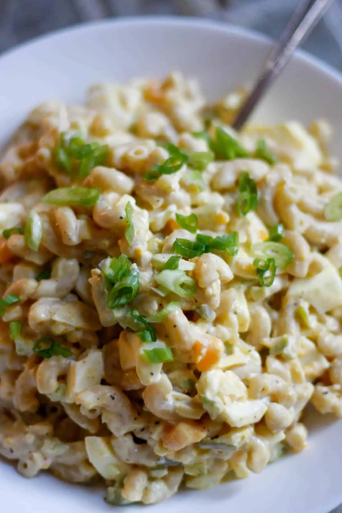 Creamy macaroni salad with chopped hard boiled eggs and green peppers garnished with green onion slices in a white bowl with the stem of a spoon pictured at the top right.