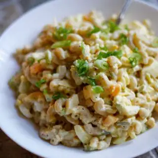 Creamy macaroni salad with chopped hard boiled eggs and green peppers garnished with green onion slices in a white bowl with the stem of a spoon in the background.