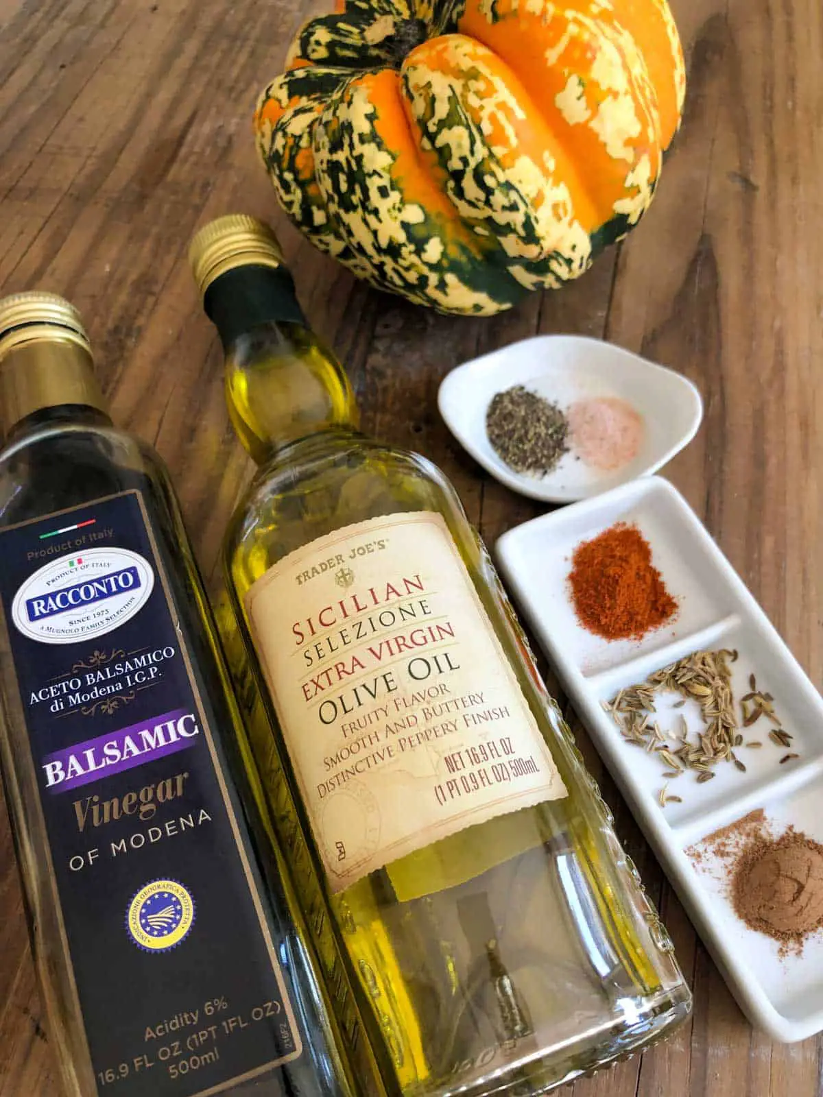 Bottle of olive oil, bottle of balsamic vinegar, a white dish with salt and pepper, a white dish with cayenne, fennel seeds and cinnamon, and a carnival squash.