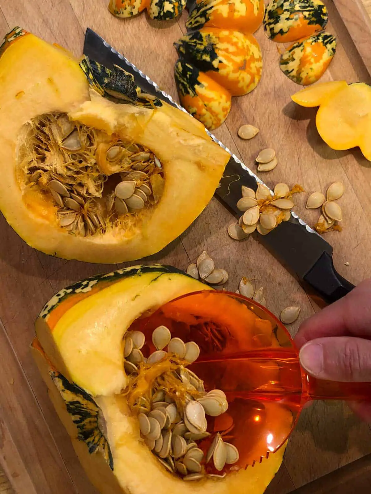 Carnival squash that has been split with a serrated knife next to it and a hand holding an orange scooper scooping out the seeds with additional seeds and cut parts of the squash laying next to the split halves.