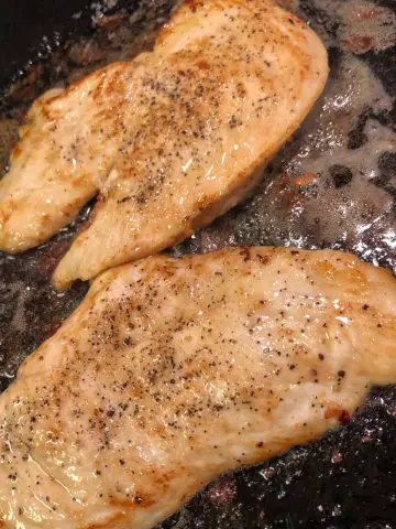 Thin slices of seasoned chicken breast cooking in a cast iron skillet in a mixture of olive oil and butter.