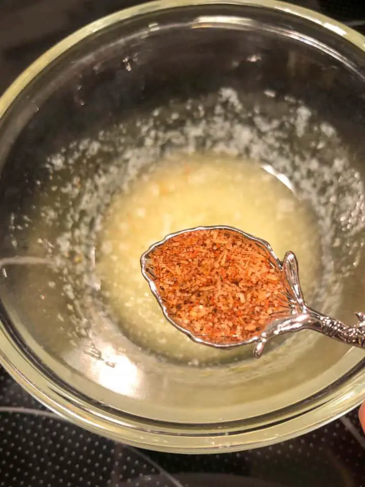 A glass bowl with melted butter in the background and a measuring spoon containing cajun spices in the foreground.
