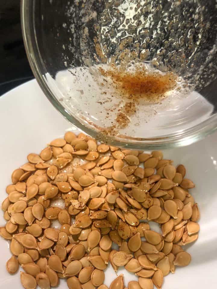Roasted Carnival Squash Seeds in a white bowl with a glass bowl containing melted butter and seasoning positioned over the seeds so that the butter can pour over them.