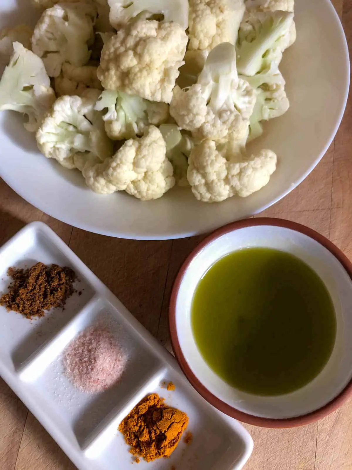 Cauliflower florets in a white bowl, olive oil in a small terracotta bowl, and garam masala, turmeric, and pink salt in a white dish.