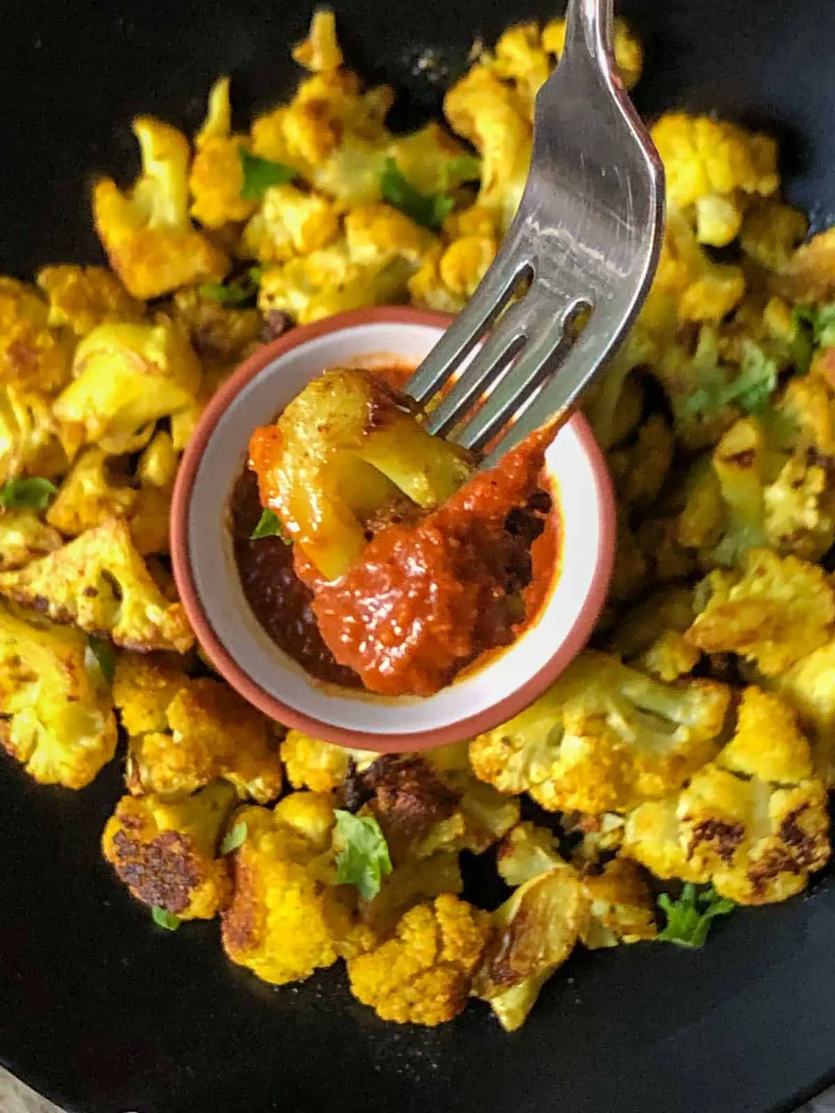 Tender Cauliflower florets roasted with Indian spices garnished with cilantro and a small terracotta bowl with red chutney and a fork piercing one of the florets which was dipped in the chutney.