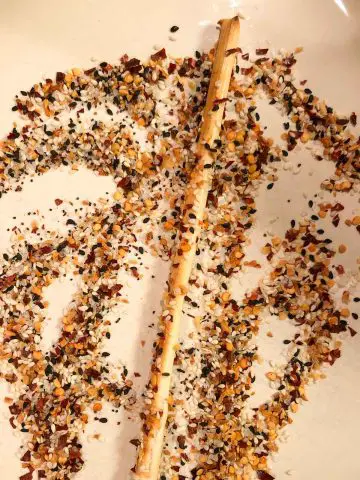 Everything bagel seasoning, salt, and red chili flakes on a dish with a thin breadstick being rolled in the seasoning so that it will coat the breadstick.