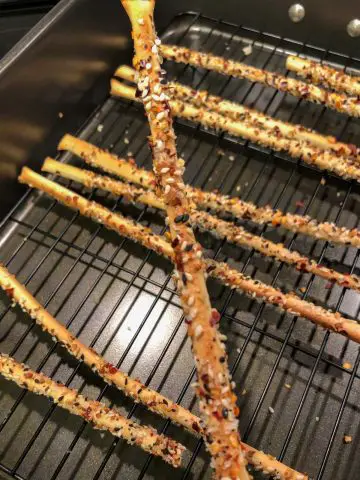 Thin breadsticks coated with everything bagel seasoning, salt and red chili flakes on a wire rack in a roasting tin with one of the breadsticks being featured in the foreground.