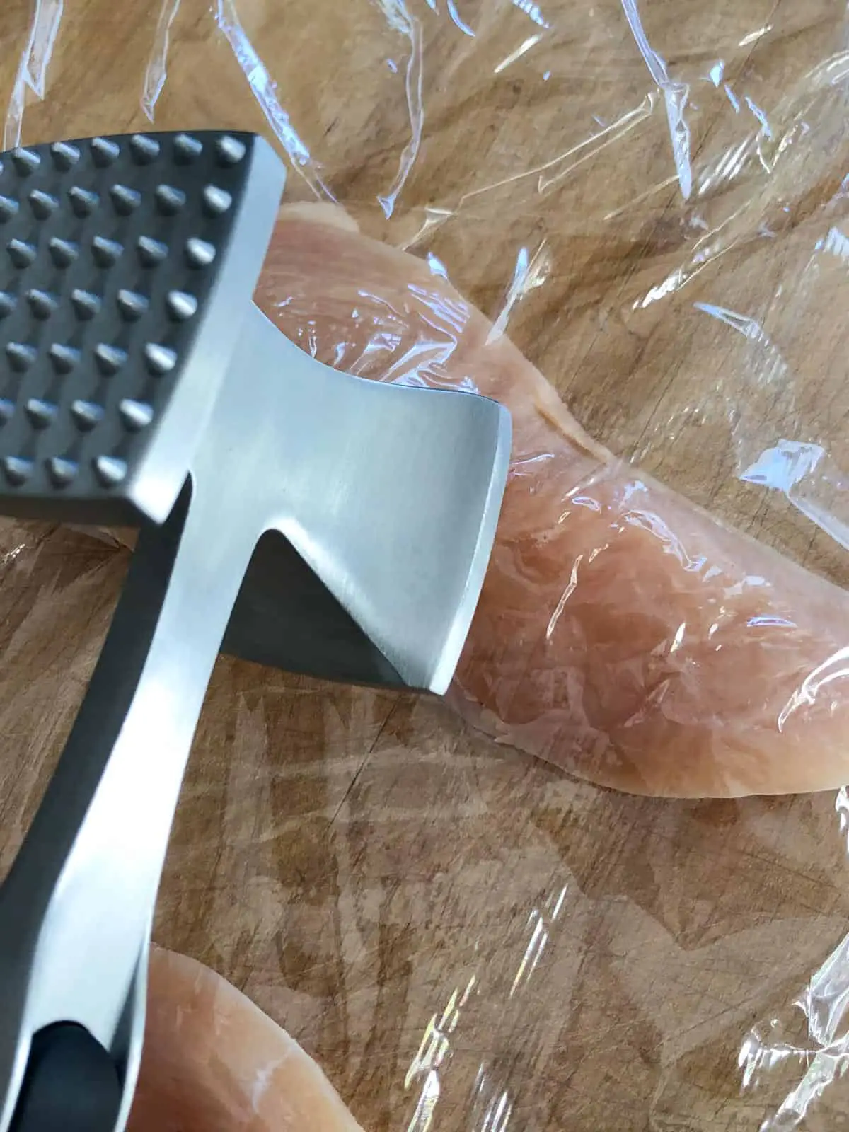 Chicken breast placed between plastic wrap with a meat mallet poised over it.