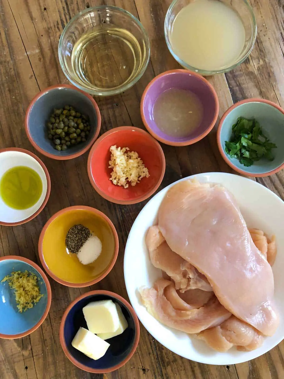 Thin slices of uncooked chicken breast in a white dish, bowls filled with butter, lemon zest, salt and pepper, olive oil, minced garlic, capers, white wine, chicken broth, lemon juice, and Italian parsley.