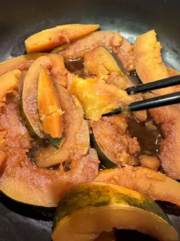 Tender wedges of acorn squash cooked in a soy braising liquid with a pair of chopsticks holding some of the squash.